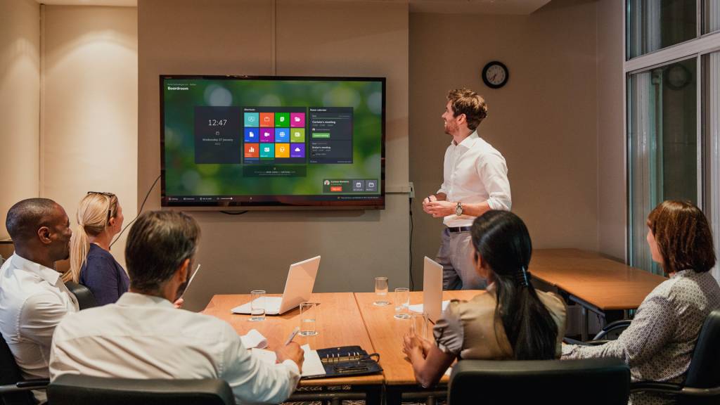 Five multi-cultural staff members facing towards a screen in a meeting room. A white man in a shirt and smart trousers is standing beside the screen, facing it. We see the Launcher interface on the screen.
