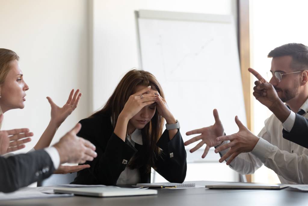 Frustrated millennial female worker sitting at table with colleagues, felling tired of working quarreling at business meeting. Upset stressed young businesswoman suffering from head ache at office.