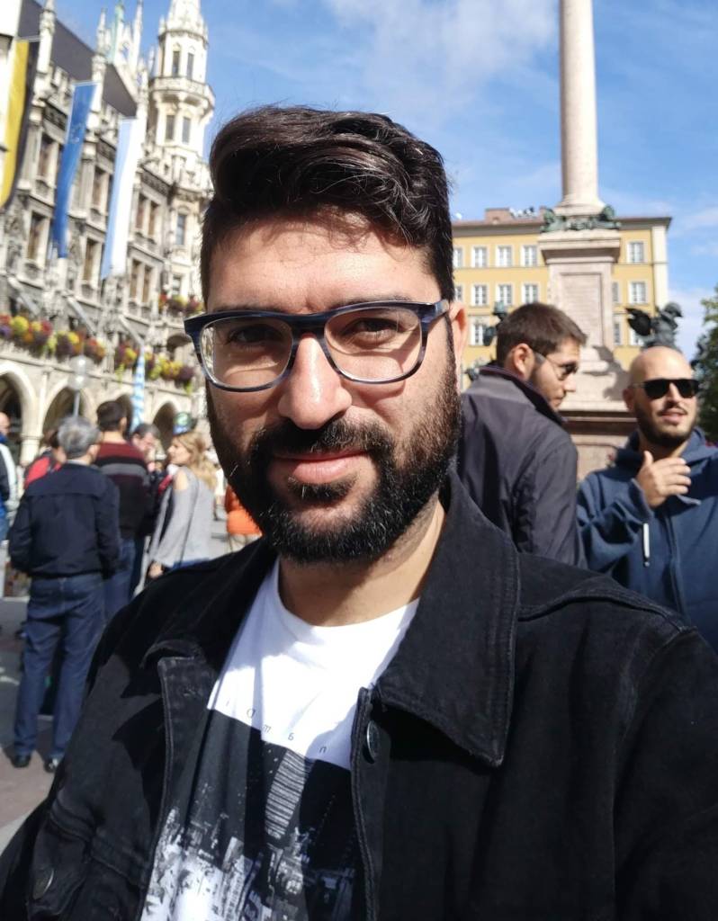 A picture of Fran. He has dark short hair, glasses and a beard. We see the city scape of a European city in the background.