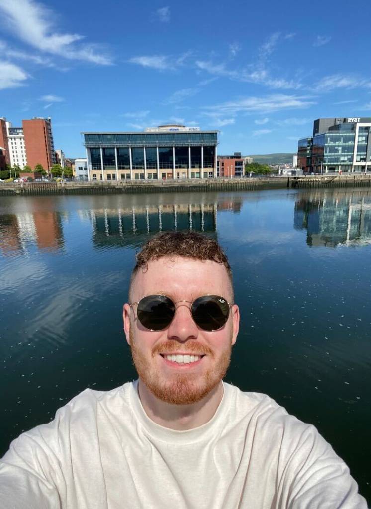 A picture of Joe taking a selfie and smiling in front of a body of water. He's wearing black sunglasses and a white t-shirt. We can see landmarks in the distance which are reflected off the surface of the water. There's a blue sky with light wisps of white cloud.