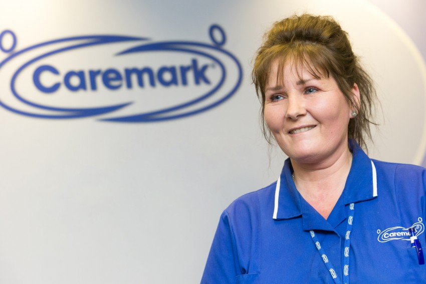 A female Caremark employee looks cheerily off into the distance. She wears a blue uniform. We see the blue Caremark logo in the background.