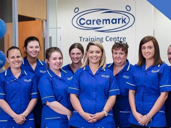 A group photo of home care staff from Caremark UK. We see 7 female staff of different ages with different hair colours in their blue Caremark uniforms standing in a line. They're smiling to the camera. They're standing in front of a wall that reads 'Caremark Training Centre'.
