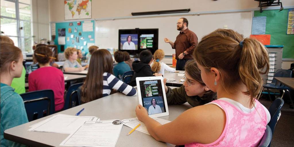 An image of a grade school classroom. We see a male teacher presenting at the front of the room while a video plays on the display at the front of the classroom. The students are attentively listening to what is being said. We also see a close up of two female students, one is holding an iPad. She is watching the same content from the interactive board on her device. 