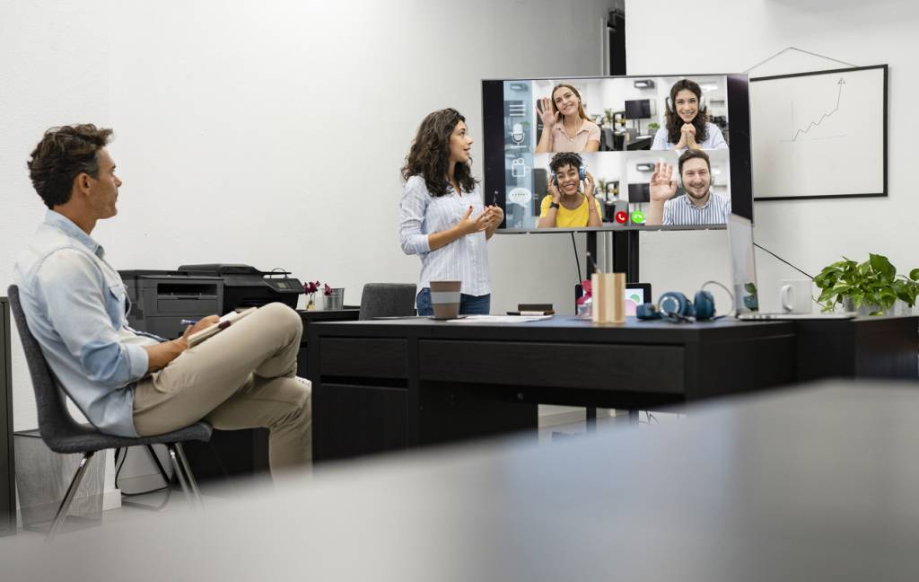 An office video call. A male and female member of the team join the video call in person from the office. They're both fancing a screen with a video call featuring four multiracial colleagues working from home