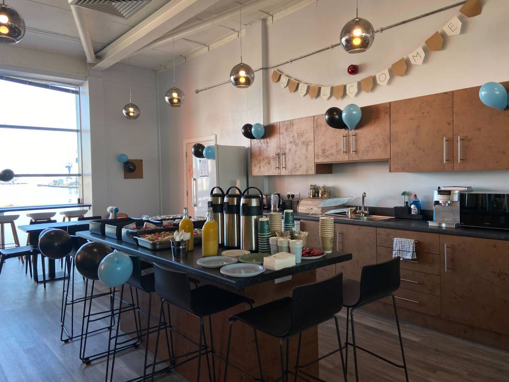 A large open-plan company kitchen. The cupboards are brown with silver chrome handles. There's an island in the middle of the image that's surrounded by high stools. The table is laid with coffee, orange juice and paper cups and plates.