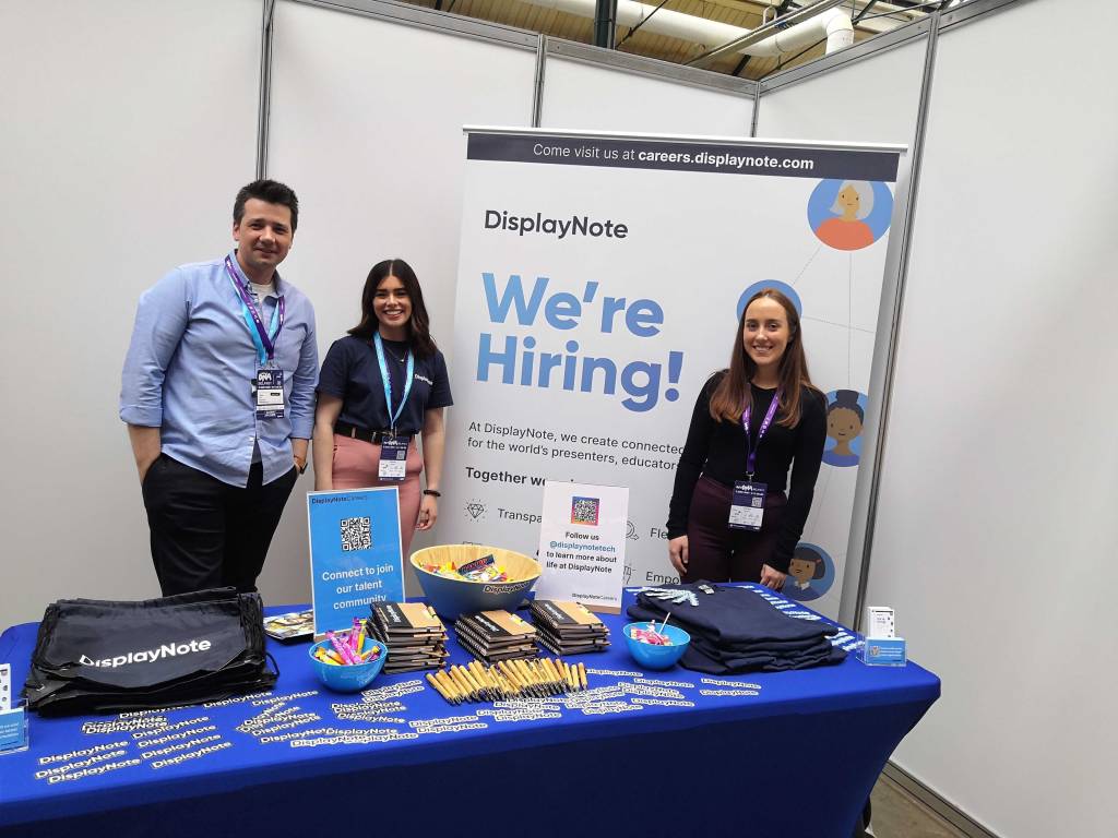 Two young women and a man in his 30's stand in front of a DisplayNote branded banner that reads 'were hiring'. They are stood behind a table with a blue table cloth. The table is covered with branded merchandise like pends, notebooks, stickers and free sweets.