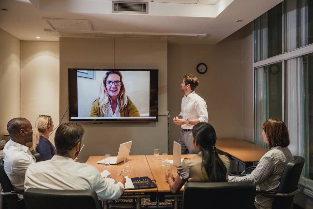 A meeting room with six different ethnicity people. The are facing away from the image towards a screen. On the screen is a video call featruing a middle aged woman with blond hair. A youngish white male is standing beside the screen. The other attendees are seated. 