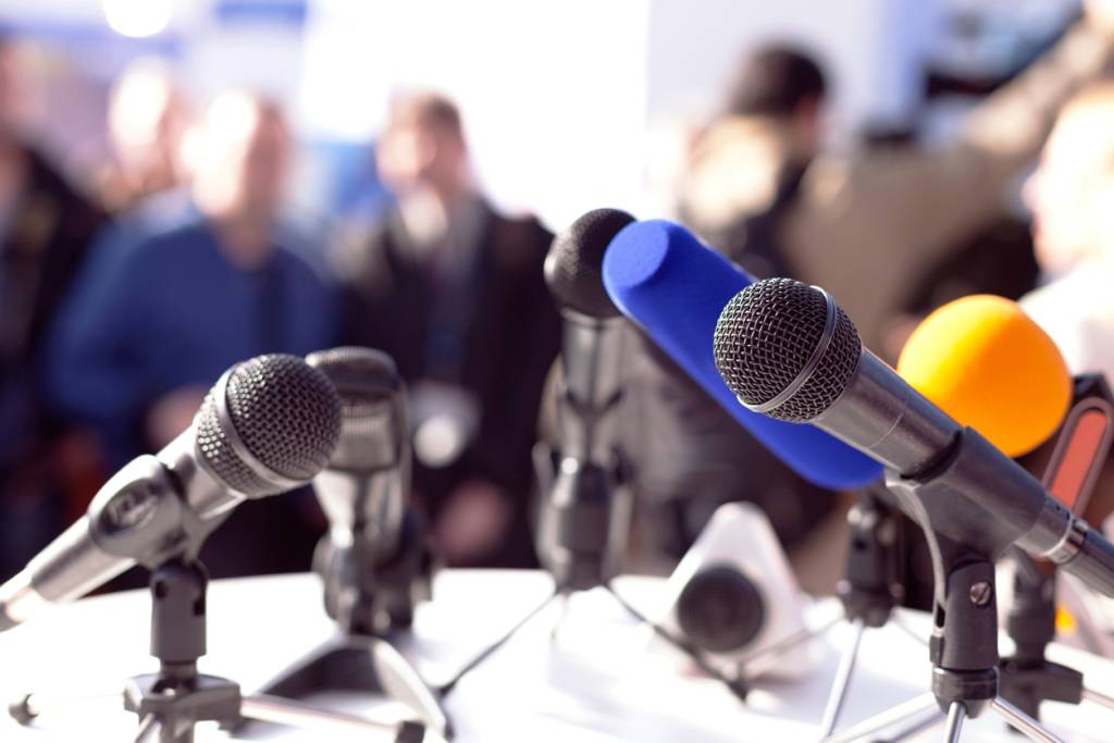 Picture of mutilple different microphones on a desk at a press conference. There is a blurred audience in the background of the image.