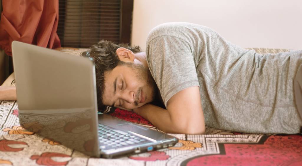 Tired young man sleeping in bed with laptop - hard work, laziness of working from home or WFH during coronavirus or covid-19 lock down concept. Difficulties of hybrid working.