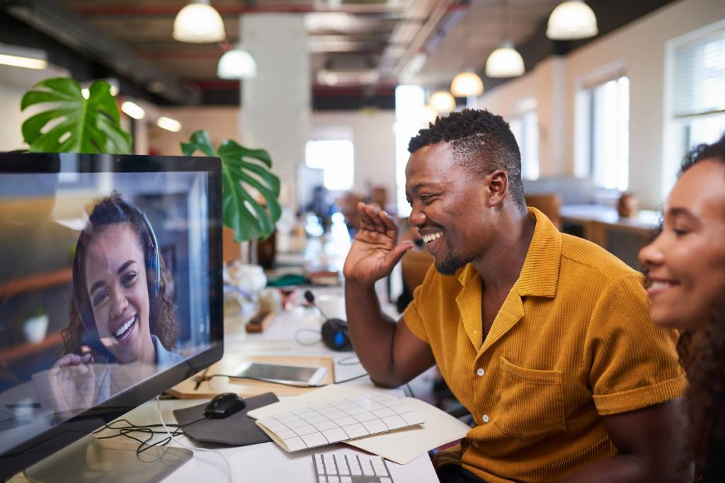 A Black man waves to his colleague on a video call from his office. Hybrid working.