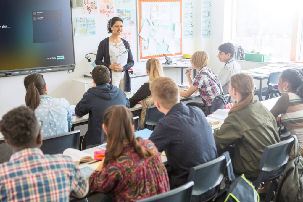 Female teacher stands at the front of the room beside an interactive screen. Students ages 15-17 are face towards the teacher in plain clothes.