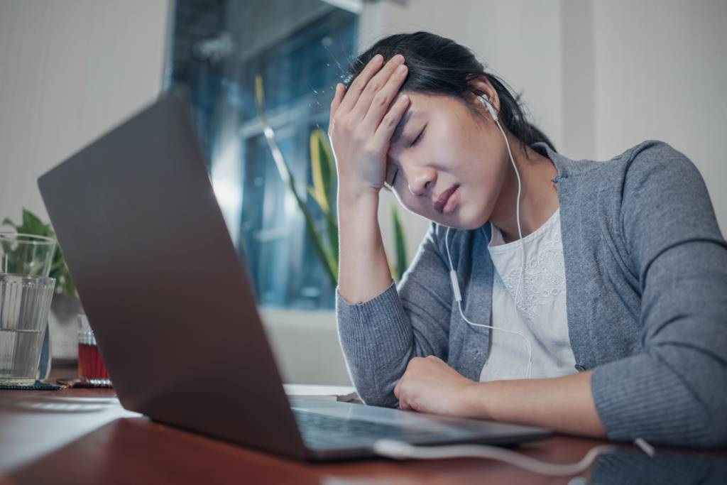 A medium shot of a stressed and frustrated young adult Asian woman, in a grey cardigan and white t-shirt inside working in home office with a laptop computer. head in hand. Bad virtual/hybrid meeting experience. 