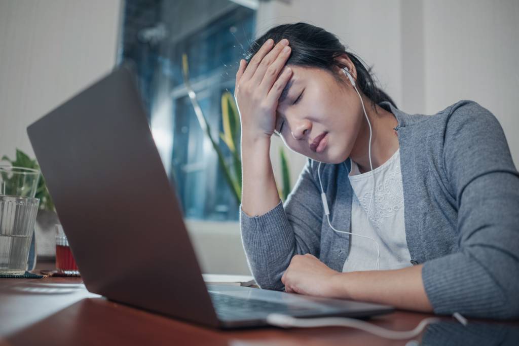 A medium shot of a stressed and frustrated young adult Asian woman, in a grey cardigan and white t-shirt inside working in home office with a laptop computer. head in hand. Bad virtual/hybrid meeting experience.