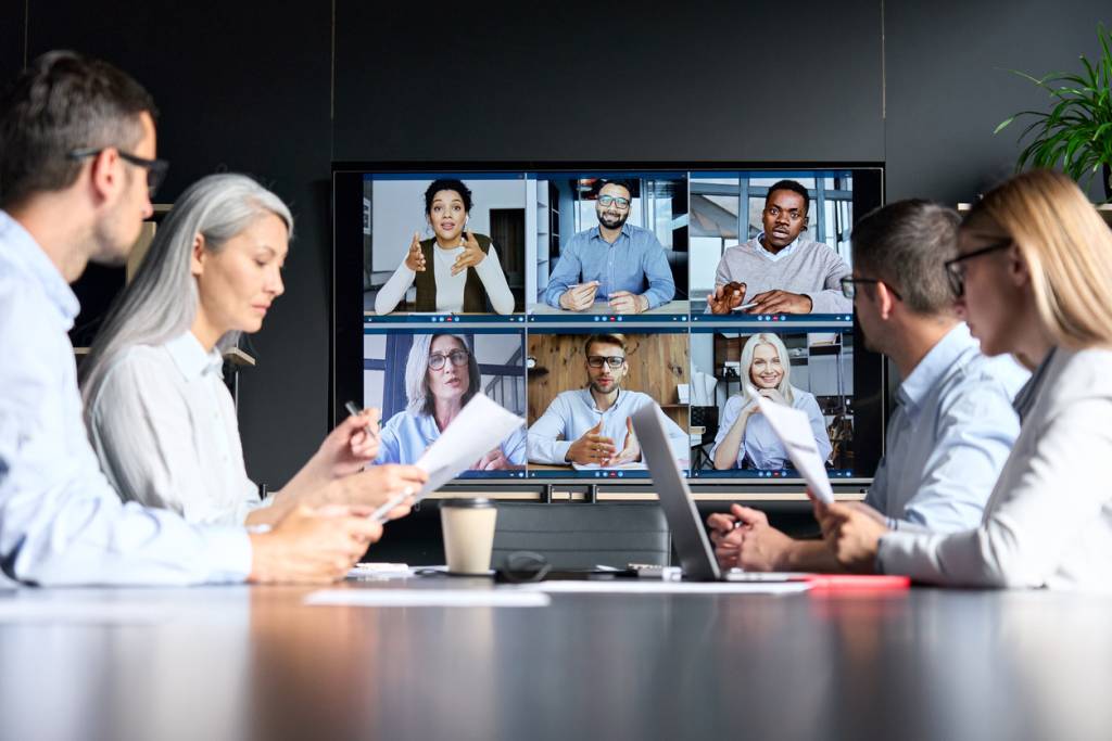 Global corporation online hybrid meeting in meeting room with diverse people sitting in modern office and multicultural multiethnic colleagues on big screen monitor. Business technologies concept.