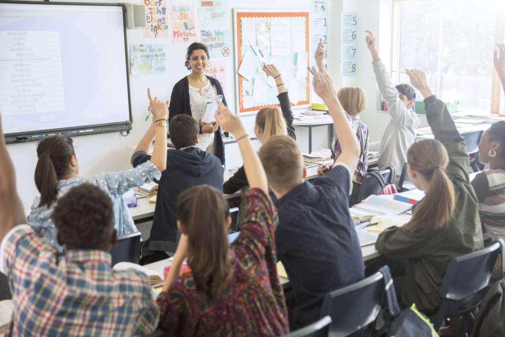 Rear view of teenage students raising hands in classroom.