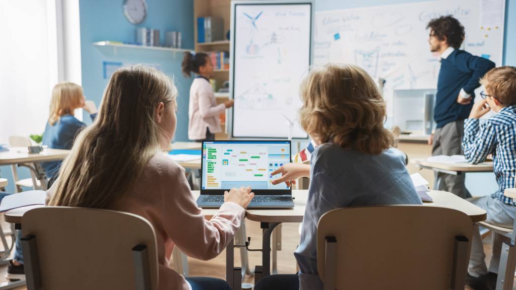 Elementary School Science Class: Over the Shoulder Little Boy and Girl Use Laptop with Screen Showing Programming Software. Physics Teacher Explains Lesson to a Diverse Class full of Smart Kids. Broadcast update.