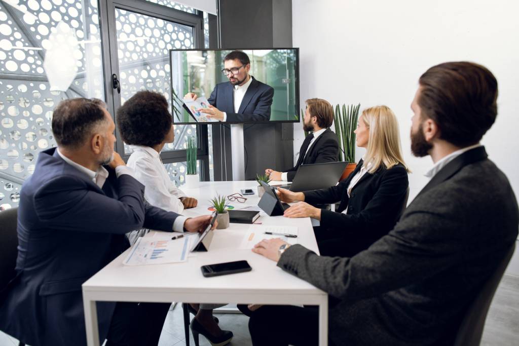 Team of five focused multiethnical businesspeople looking at screen, having video conference in modern office, with their male colleague explaining some financial charts. Telemeeting.