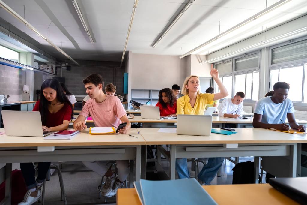 Teenagers in a classroom using their laptop. There is a girl raising her hand to ask a question